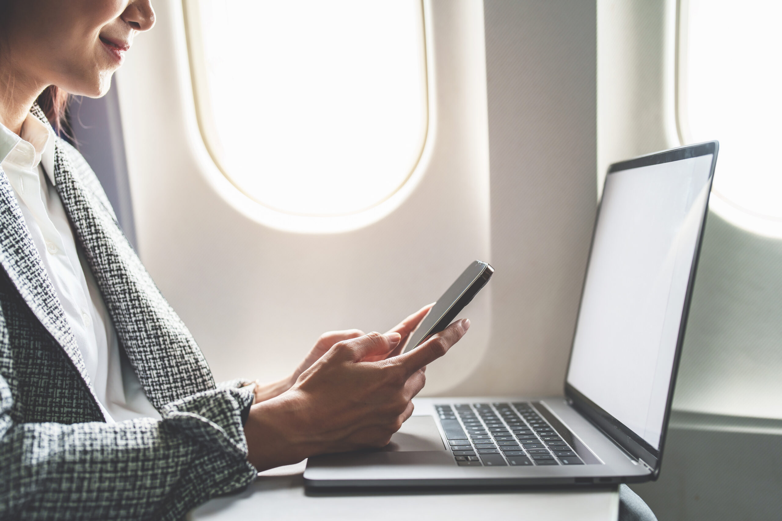 A successful asian businesswoman or female entrepreneur in formal suit in a plane sits in a business class's seat using a smartphone and computer laptop during flight